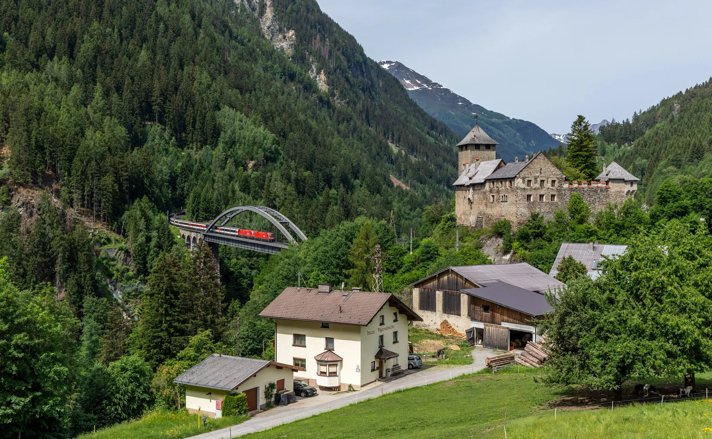 A red locomotive crossing the arched Trisanna bridge with Wiesberg castle in front of forested slopes.