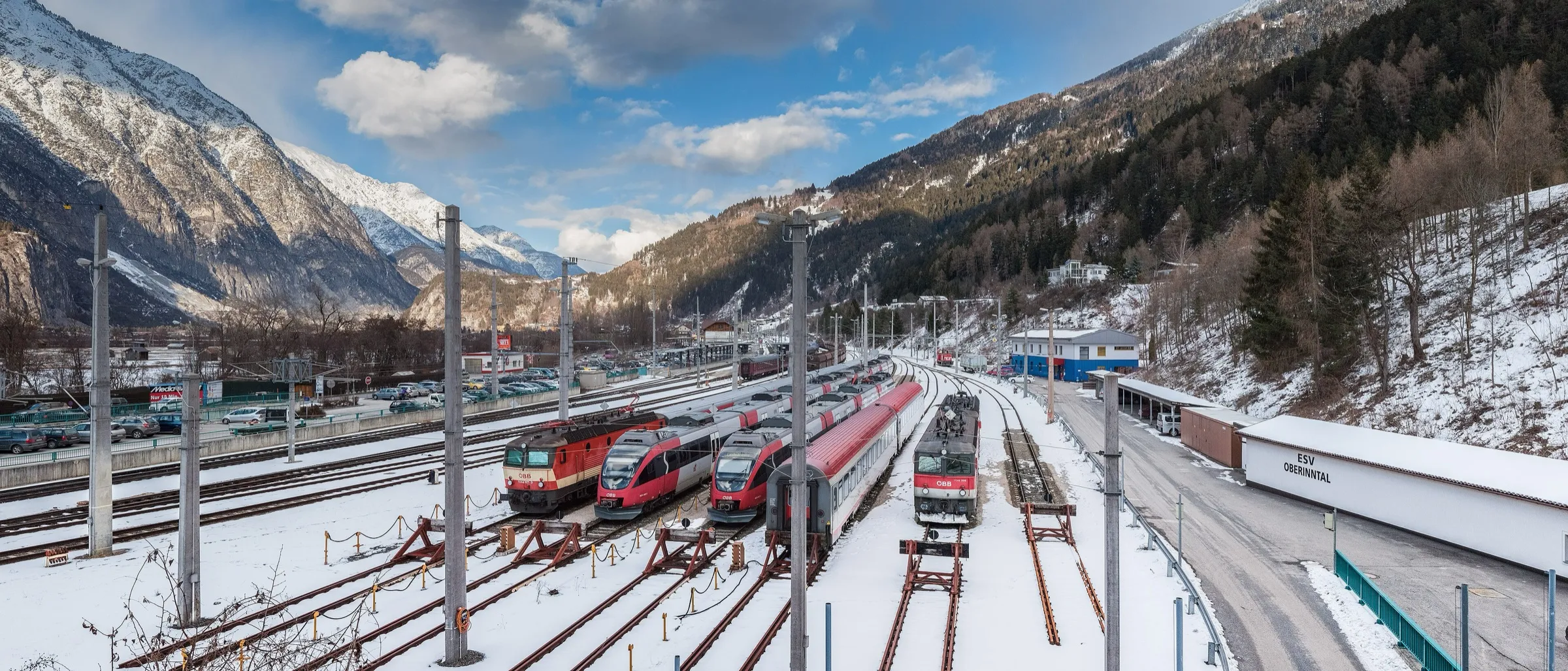 A wide winter view of Landeck-Zams station with red ÖBB trains in the sidings and snowy mountains all around.